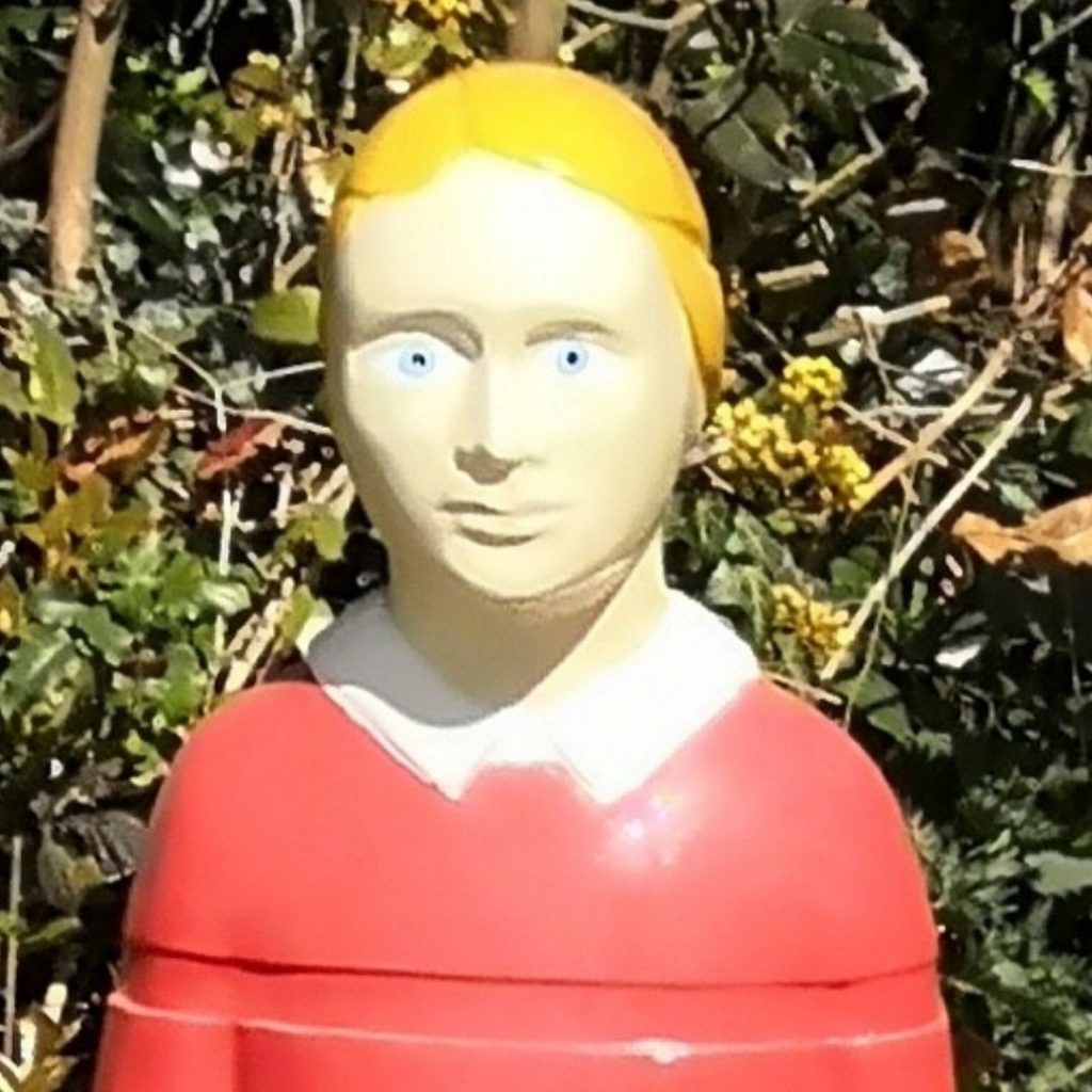 A closer cropped version of the statue in the photo above. This version just shows the statue from the shoulders up, so you can see bright red plastic jumper, with painted white collar, and a startling face. the statue has no ears, and has painted buttercup yellow hair. The eyes are very bright blue, and the facial expression is difficult to read, looking curiously blank, with perhaps a slight hint of the a smile. The lips and eyebrows are the same colour as the skin - a sort of off white, that gives it a slight hint of corpse.