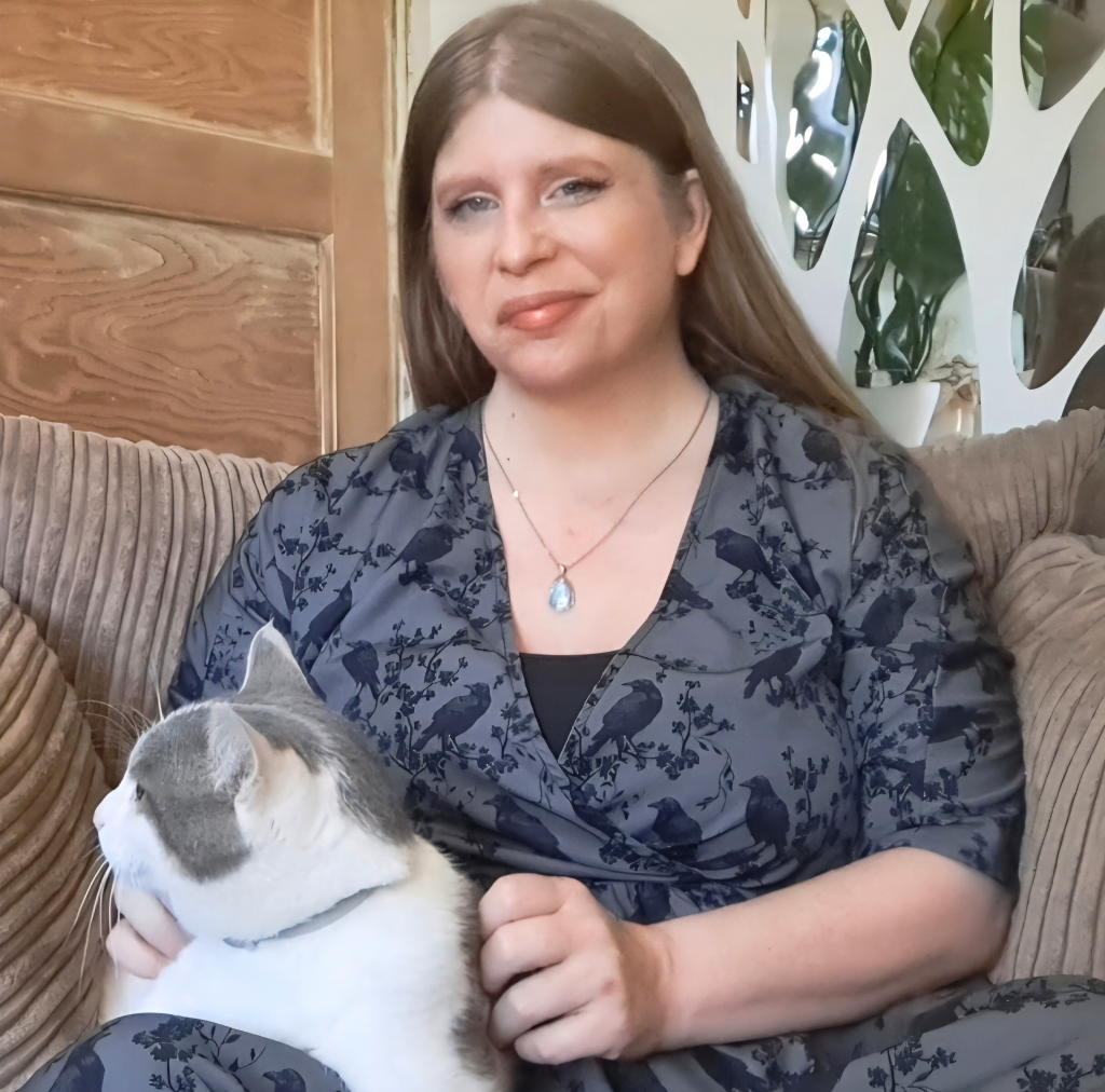 V, the creator of The Living Craft, Building your personal practice, is a 40 year old white woman with long blonde hair. She is sitting on a comfy, soft armchair, stroking a white cat with grey smudges on him. She is wearing a grey wrap dress with a subtle raven pattern on and smiling warmly at the camera.